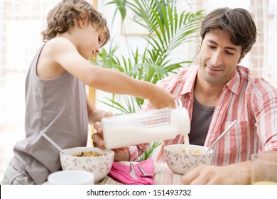 Single Parent Dad And His Young Son Having Breakfast Together And Eating Cereals With The Boy Helping And Pouring Milk In His Father Cereal Bowl During A Sunny Morning At Home.