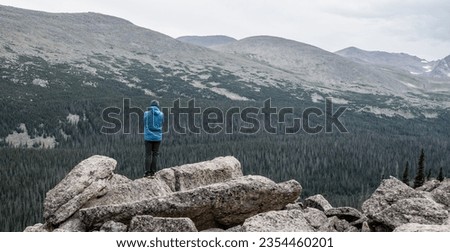 Single Person In Blue Coat Stands On Rocks overlooking Stormy Peaks Pass