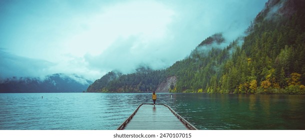A single person walks along a wooden dock stretching out into a still lake. The mountains rise up in the background, shrouded in mist, creating a tranquil and atmospheric scene. - Powered by Shutterstock