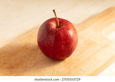 Single Red Apple with Stem Resting on a Light Wooden Cutting Board in Soft, Warm Light