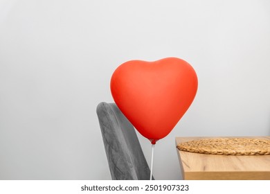 A single red heart-shaped balloon is attached to the back of a grey chair, which is positioned beside a wooden table with a woven placemat. The setting appears to be indoors, with a minimalistic. - Powered by Shutterstock