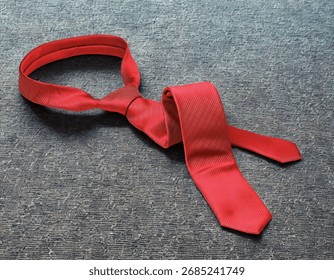 A single red necktie with a subtle pattern is casually draped on a dark, textured background, suggesting a formal accessory.