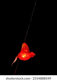 A single, red paper lantern in the shape of a bird hangs against a dark background, illuminated from within, creating a warm and inviting glow.