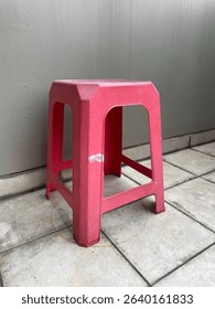 A single red plastic stool is placed outdoors on a tiled floor against a plain grey wall