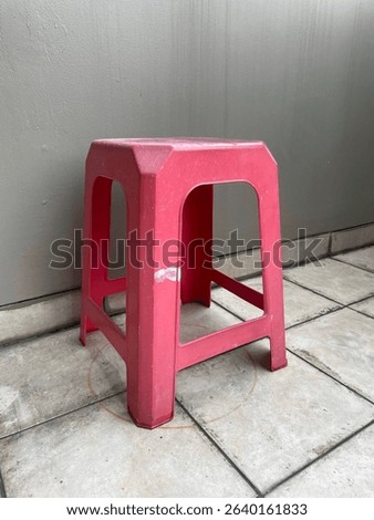 A single red plastic stool is placed outdoors on a tiled floor against a plain grey wall