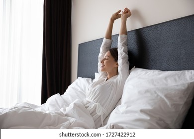 Single rested renewed after deep healthy night sleeping 30s woman wake up do stretching exercises seated in bed, satisfied female raised hands closed eyes enjoy early morning at home hotel room alone