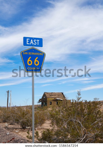 A\
single road sign marks historic route 66 going East through San\
Bernardino County, California. A single isolated derelict wooden\
cottage stands in the background under a big blue\
sky.