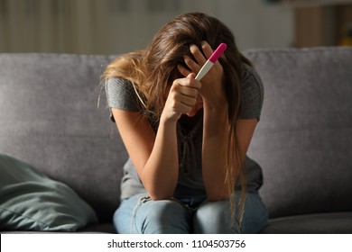 Single sad woman complaining holding a pregnancy test sitting on a couch in the living room at home