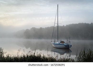 A single sailboat on a lake during a foggy autumn sunrise. In the background presented lake shore with forest barely visible due to dense fog.  - Powered by Shutterstock