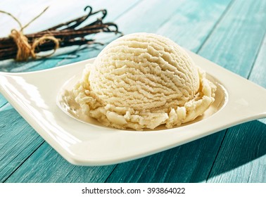 Single scoop of creamy vanilla ice cream on a stylish modern plate on a blue wooden picnic table outdoors in summer with a bundle of dried vanilla pods behind
