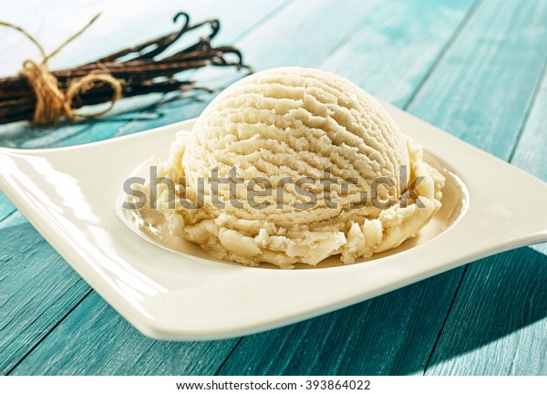 Single scoop of creamy vanilla ice\
cream on a stylish modern plate on a blue wooden picnic table\
outdoors in summer with a bundle of dried vanilla pods\
behind