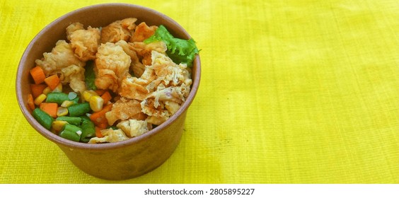 A single serving of rice packaged in a convenient brown paper bowl. Atop the white rice sits a golden-brown crispy fried chicken topping, slices of fried omelet, and a vegetable mix of green beans, carrots, and sweet corn. A fresh lettuce leaf adds a splash of color to the dish. The rice bowl is presented on a bright yellow surface under natural light, evoking the image of a delicious, nutritious, ready-to-eat meal—perfect for lunch, catering, or a packed meal.


