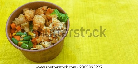 A single serving of rice packaged in a convenient brown paper bowl. Atop the white rice sits a golden-brown crispy fried chicken topping, slices of fried omelet, and a vegetable mix of green beans, carrots, and sweet corn. A fresh lettuce leaf adds a splash of color to the dish. The rice bowl is presented on a bright yellow surface under natural light, evoking the image of a delicious, nutritious, ready-to-eat meal—perfect for lunch, catering, or a packed meal.


