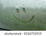 A single, simple smiley face is drawn on a car window. The window is covered in raindrops, obscuring the view outside. The smiley face is visible through the water droplets