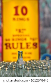 Single Soda Pop Bottle In Ring Toss Carnival Amusement Park Game Detail Above Many Glass Bottles With Yellow And Red Rules Sign Blurred In Background. Fairgrounds Close Up