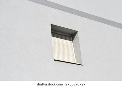 A single square window with light beige closed roller shutters is recessed into a plain white textured building wall with a diagonal shadow