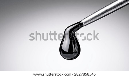 A single, thick drop of dark, viscous liquid, possibly oil or tar, is suspended from the end of a metallic instrument, against a neutral background.
