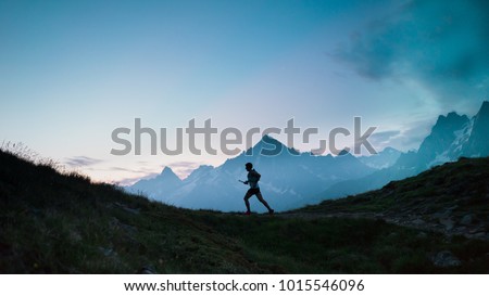 A single trail runner, silhouetted against the sky at sunrise while running in the mountains of the Alps along a steep trail