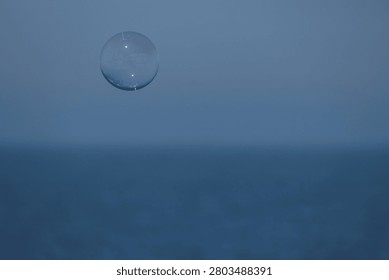 A single translucent soap bubble floats against a seamless blue-grey misty seascape where the sky and sea merge into one soft gradient background. The bubble sits in the upper left of the frame, its surface showing a subtle iridescent sheen and tiny light reflection. The vast empty blue-grey space creates enormous negative space ideal for text overlay. Minimalist, conceptual, and emotionally evocative — communicating fragility, dreams, impermanence, childhood, and freedom. Ideal for editorial, advertising, book covers, wellness, mental health, and conceptual art use. No people, full commercial license.