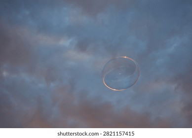 Single translucent soap bubble reflecting subtle pink, blue and green colors against a cloudy evening sky in Zadar, Croatia.