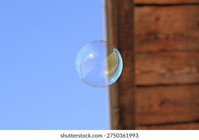 Single transparent soap bubble floating in the air with iridescent blue, yellow and pink reflections, photographed against a bright blue sky and a wooden structure background.
