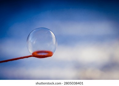 A single transparent soap bubble rests delicately on an orange plastic bubble wand, set against a softly blurred, ethereal background of blue sky and light clouds, capturing a fleeting moment of quiet, playful wonder.