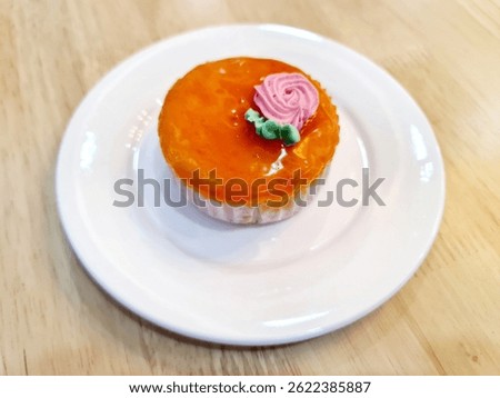 A single, vibrant mini mango cheesecake sits centered on a clean white plate, placed on a light-toned wooden surface. The individual dessert features a smooth, bright orange mango topping 