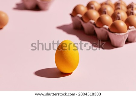 A single, vibrant yellow egg is placed in front of a carton filled with brown eggs, set against a soft pink surface with dramatic shadows.