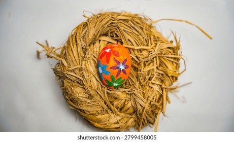 A single, vibrantly painted Easter egg rests in a rustic straw nest against a clean white backdrop.