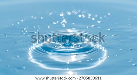 A single water drop is captured mid-fall, creating concentric ripples on a smooth, bright blue liquid surface. Numerous small, bright bokeh lights are scattered across the surface.