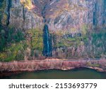 A single waterfall moving over blacken rock with white foaming water flows down the side of a moss laden cliff surrounded by barren trees in the Fall on the edge of the Columbia River in the Columbia 