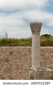 A single weathered column with a simple cushion capital stands before a drystone wall in the Palace of Gordian at Volubilis, with a green grassy background and clear sky overhead.