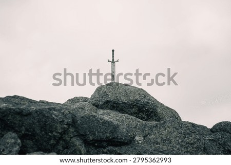 A single weathered medieval-style sword is driven upright into a cluster of dark granite rocks on a barren hilltop. The steel blade shows signs of age