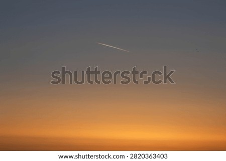 Single white inversion trail from an airplane in the sky. Abstract blurred gradient sunrise or sunset background with warm orange and gray tones. Multicolored sky with copyspace.