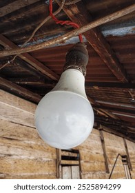 A single, white LED light bulb hangs from a wooden ceiling by a red wire and a vintage socket. The light bulb is slightly dirty and the surroundings appear rustic.