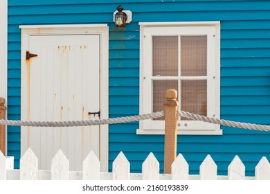 A single white panel wooden door with a vintage metal latch handle in a blue building with a single glass casement window. There's a white picket fence and rope banister in front of the entranceway. 