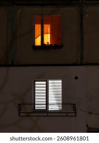 A single window emits warm orange light while another below remains dark and unlit, showcasing the contrast between vibrant life and quiet solitude in an urban setting at night.