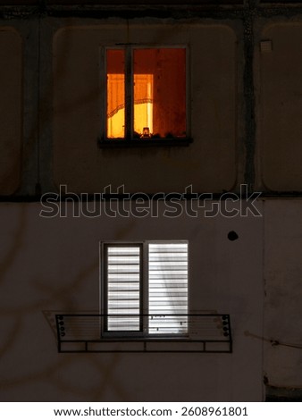 A single window emits warm orange light while another below remains dark and unlit, showcasing the contrast between vibrant life and quiet solitude in an urban setting at night.