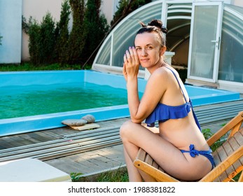 A Single Woman In A Bathing Suit Is Sitting On A Sun Lounger In The Backyard By The Pool.