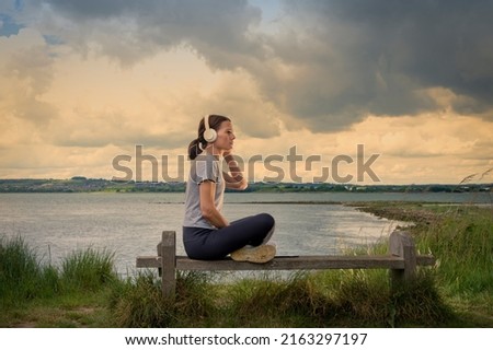 Single woman sitting on a bench beside water listening to music on her headphones. Stock photo © 