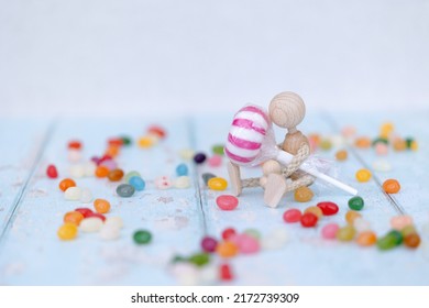 A Single Wooden Mannequin Holding A Red And White Lollipop, Standing And Sitting, On Blue Wooden Background Surrounded By Colourful Jellybeans, Space For Text 