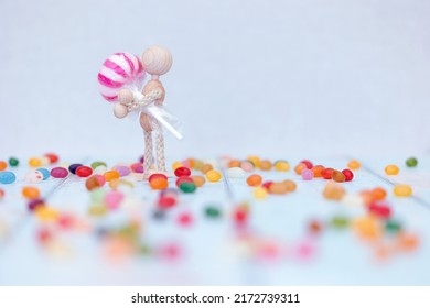 A Single Wooden Mannequin Holding A Red And White Lollipop, Standing And Sitting, On Blue Wooden Background Surrounded By Colourful Jellybeans, Space For Text 