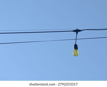 A single yellow string light bulb hanging on a wire against a clear blue sky background.