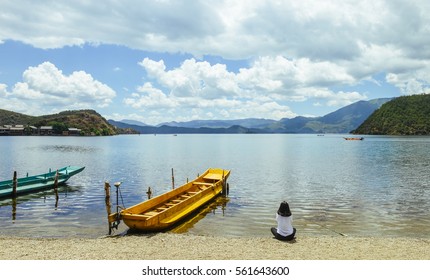 Single Young Chinese Woman Looks Into Skyline, Sitting On Bank Of Lugu Lake, Lijiang, China, Back To Camera