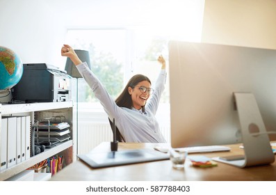 Single young excited woman celebrating something with both arms extended upward while seated at desk in small office