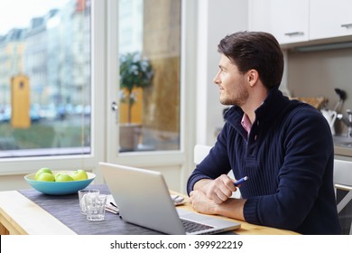 Single Young Man Wearing Blue Collar Sweater Seated With Laptop On Table In Kitchen Looking Out Window