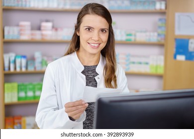 Single Young Pharmacy Technician In Long Brown Hair And White Lab Coat Entering Prescription Order On Computer With Medications On Shelf In Background