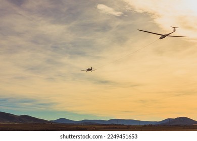 A single-engine aircraft tows a glider against the backdrop of mountains at sunset.