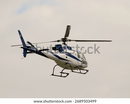 A single-engine, blue and white helicopter with visible rotor blur is captured in flight against a mostly white, cloudy sky. High quality photo