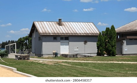 A single-story concrete utility building featuring a weathered metal roof and white double doors sits on a green lawn beside a concrete path and small arborvitae trees under a cloudy blue sky